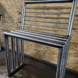 Prototyp krzesła z polerowanych rur stalowych, spawany w warsztacie na tle płyty OSB z odręcznymi notatkami.