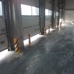 Wnętrze hali produkcyjnej z widocznymi segmentowymi bramami przemysłowymi z oknami, zabezpieczonymi żółto-czarnymi słupkami ostrzegawczymi.