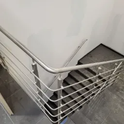 Metalowa balustrada schodowa z poziomymi wypełnieniami, widok z góry na ciemne schody i szarą podłogę z widocznymi zabrudzeniami po remoncie.