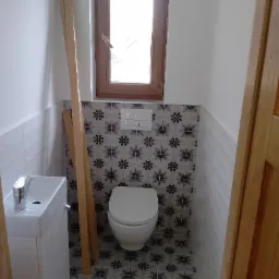 Mała łazienka po remoncie z umywalką, toaletą i drewnianym oknem, wykończona płytkami wzorzystymi na podłodze i ścianie za toaletą oraz białymi kafelkami po bokach, z drewnianymi listwami opartymi...