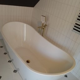 AnmarBud - Wolnostojąca wanna w nowoczesnej łazience z białymi, pionowo ułożonymi płytkami i czarnym sufitem, złota bateria wannowa z prysznicem.