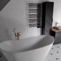 Nowoczesna łazienka z wolnostojącą białą wanną, czarnym grzejnikiem drabinkowym, szafką umywalkową z drewnianym blatem i złotą armaturą, wykończona białymi płytkami i czarnymi akcentami.