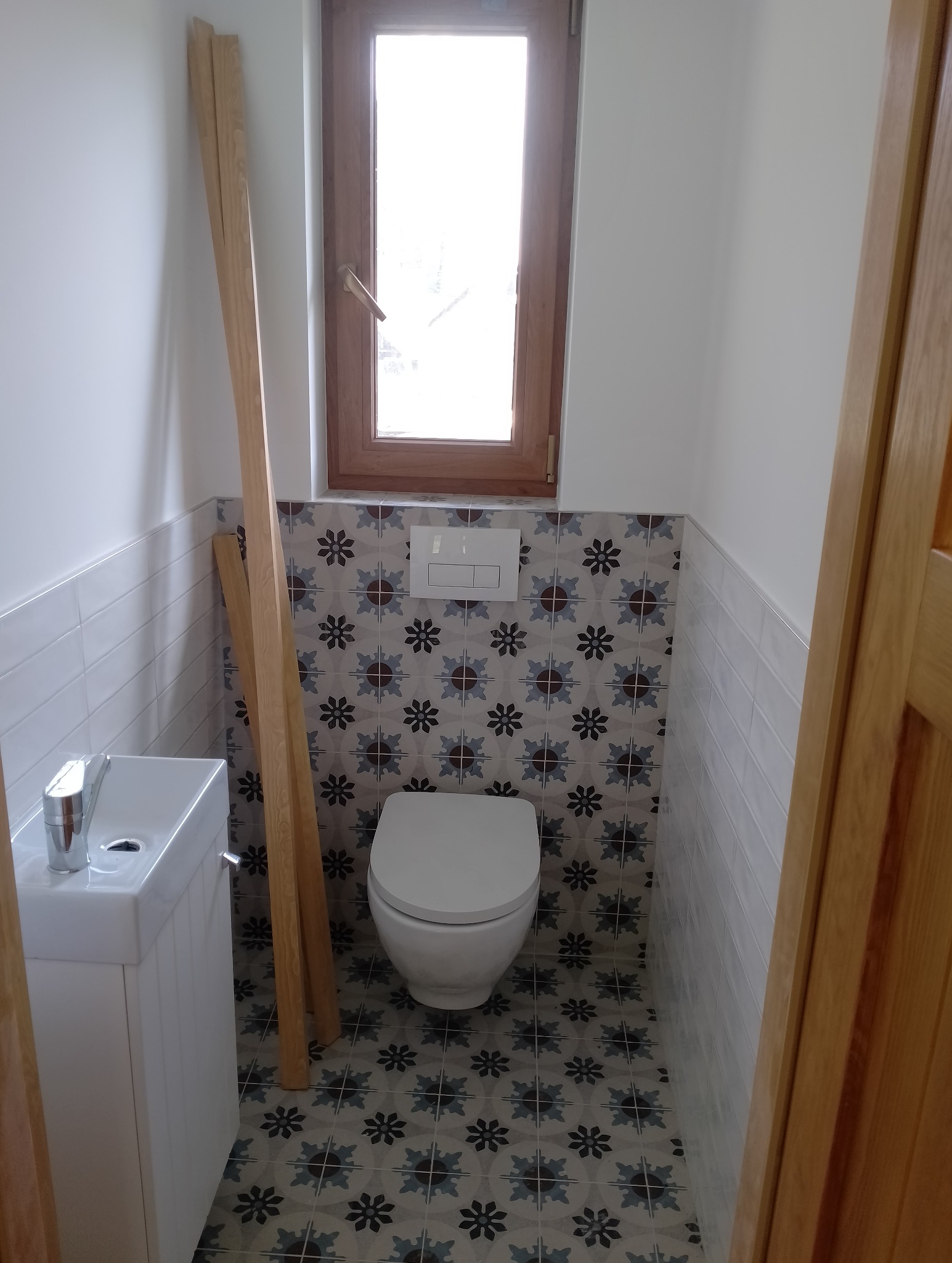 Mała łazienka po remoncie z umywalką, toaletą i drewnianym oknem, wykończona płytkami wzorzystymi na podłodze i ścianie za toaletą oraz białymi kafelkami po bokach, z drewnianymi listwami opartymi...