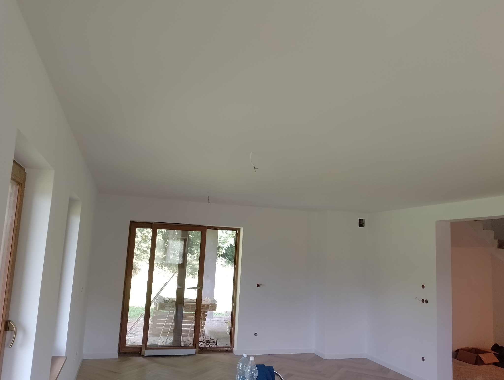 Jasne, przestronne wnętrze po zakończonym malowaniu ścian i sufitu na biało, widoczne gniazdka elektryczne, parkiet ułożony w jodełkę, przeszklone drzwi przesuwne z widokiem na zieleń.