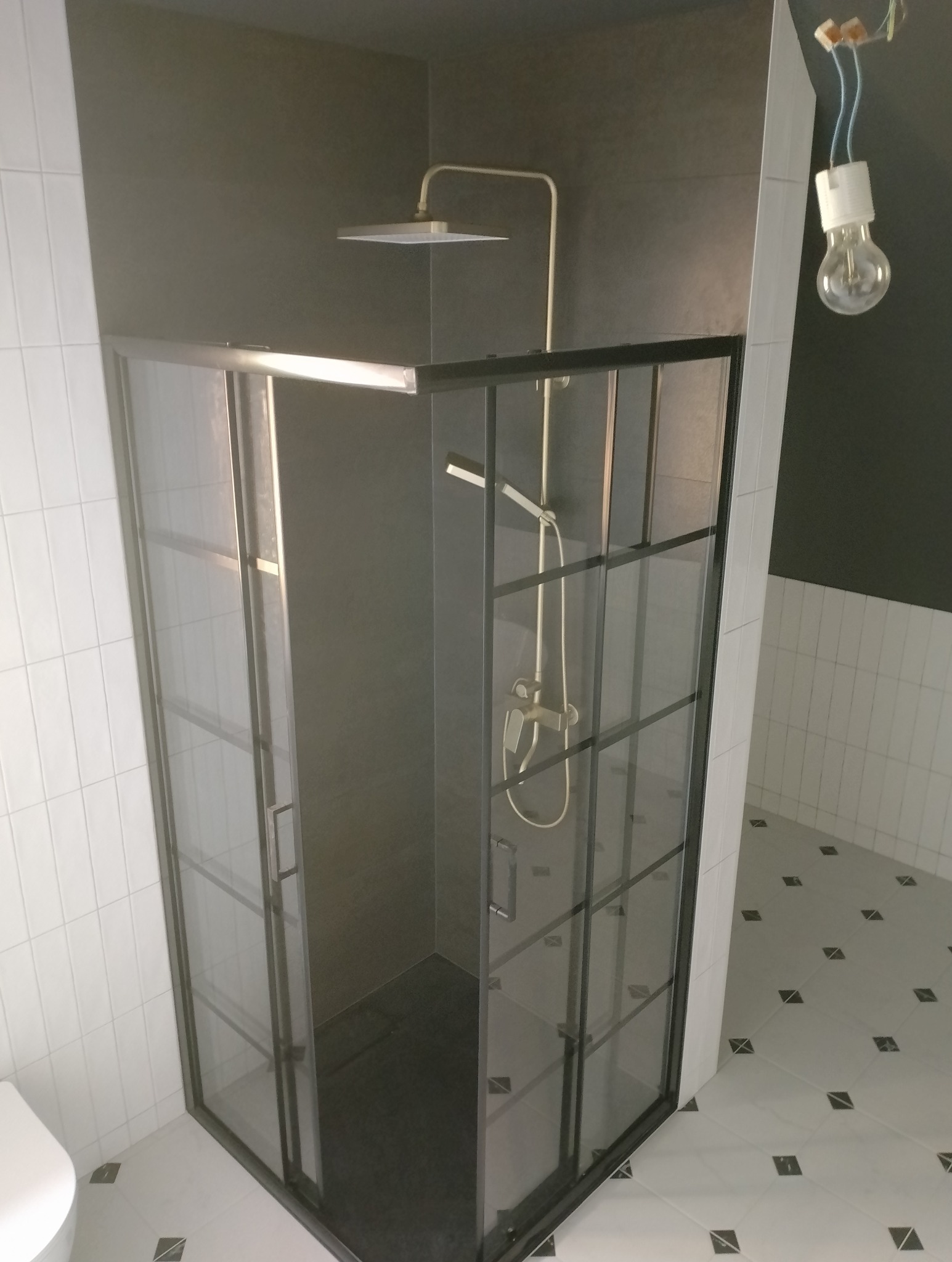 Nowoczesna kabina prysznicowa z czarnymi profilami i deszczownicą w wykończonej łazience z biało-czarną podłogą i niedokończonym oświetleniem.