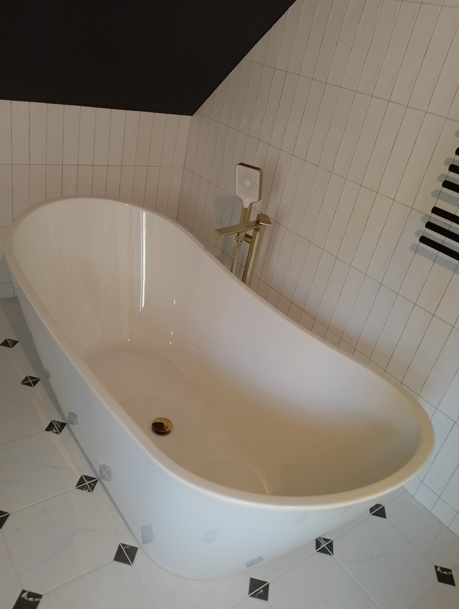 Wolnostojąca wanna w nowoczesnej łazience z białymi, pionowo ułożonymi płytkami i czarnym sufitem, złota bateria wannowa z prysznicem.