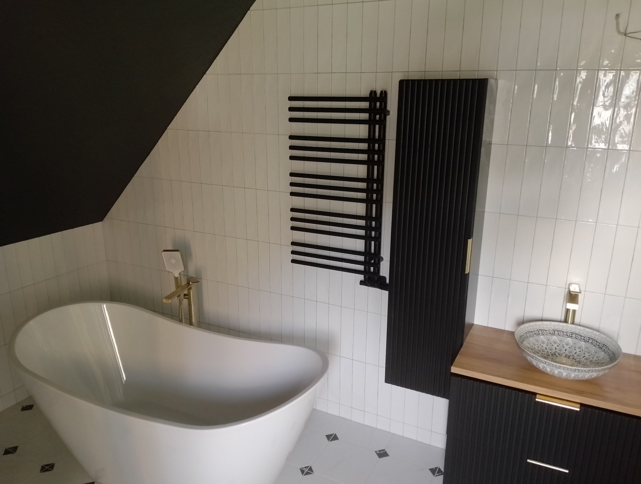 Nowoczesna łazienka z wolnostojącą białą wanną, czarnymi grzejnikami drabinkowymi i umywalką nablatową w stylu boho, na tle białych, pionowo ułożonych płytek i czarnego skosu sufitu.