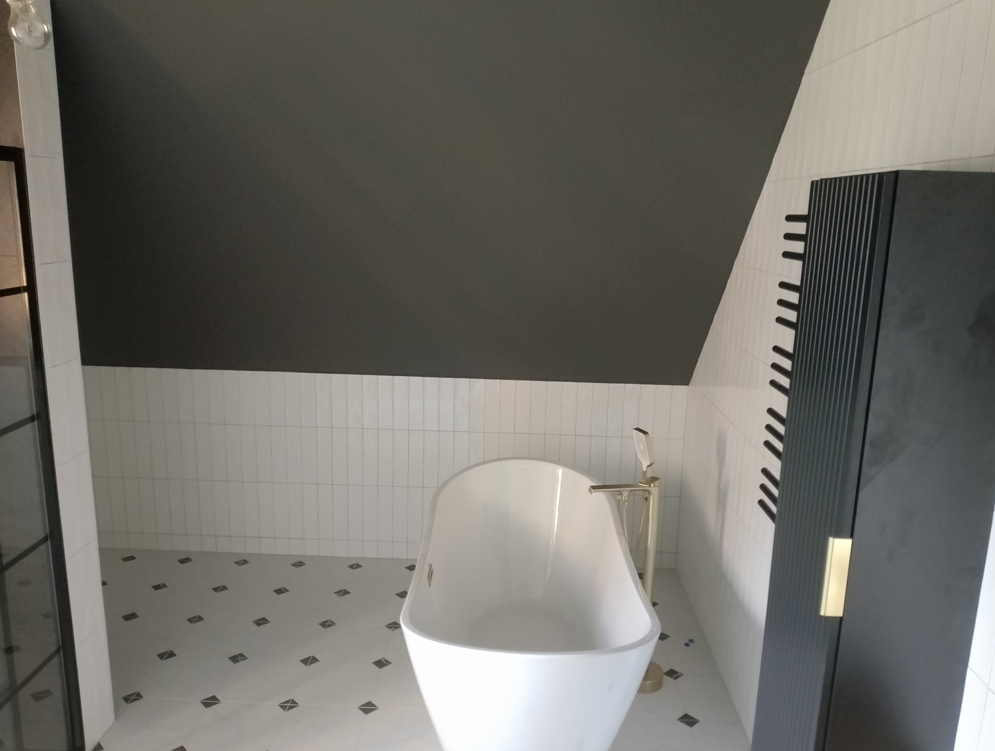 Nowoczesna łazienka na poddaszu z wolnostojącą białą wanną, złotą armaturą, czarnym grzejnikiem drabinkowym i biało-czarną podłogą w geometryczne wzory.