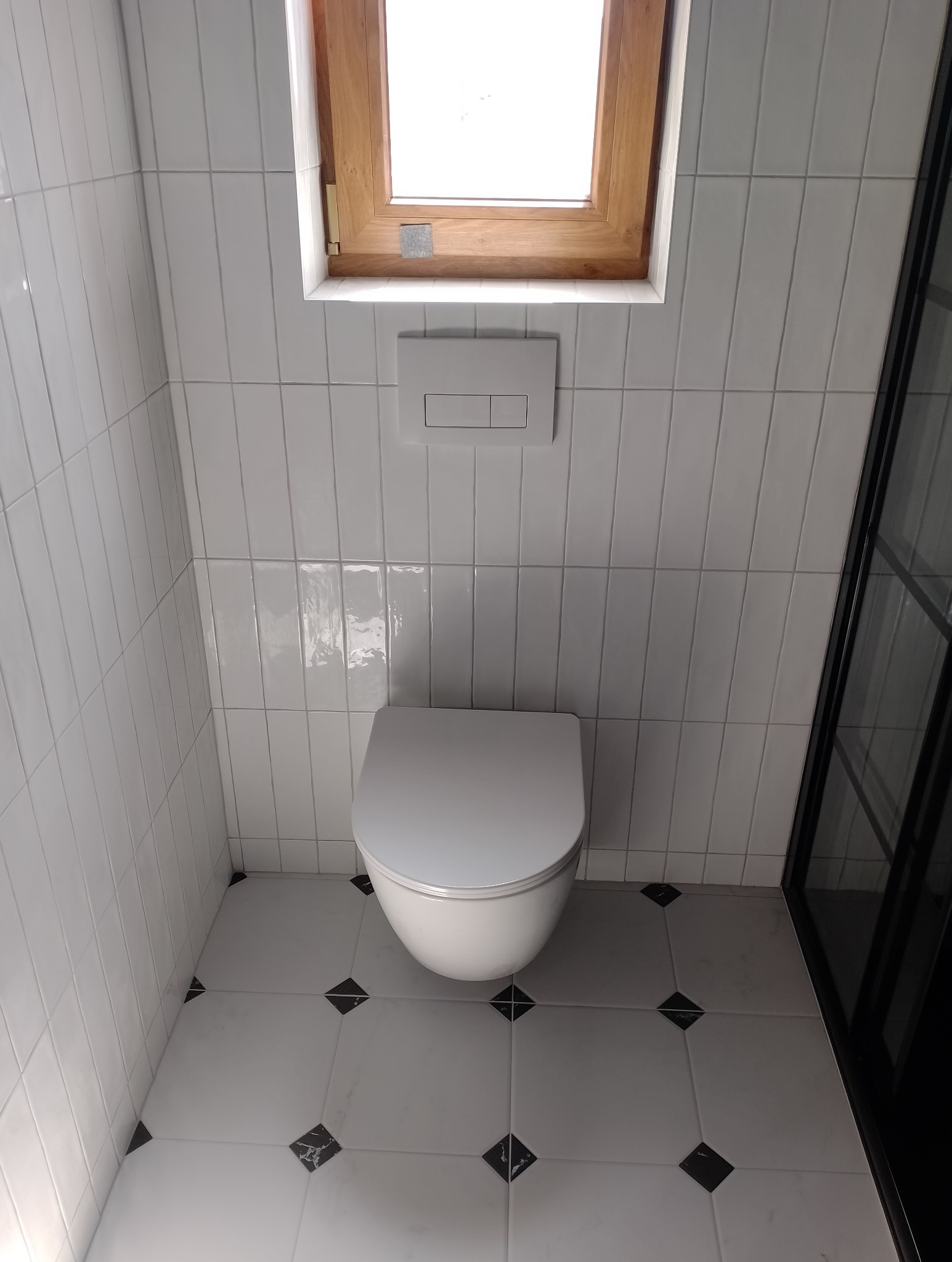 Nowoczesna toaleta wisząca w łazience wykończonej białymi, pionowymi płytkami i podłogą z białych płytek z czarnymi akcentami, oświetlona przez okno dachowe w drewnianej ramie.