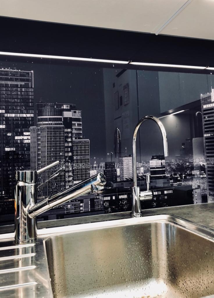 Bateria kuchenna z dwoma kranami, chromowana, na tle panelu z motywem panoramy miasta nocą, widoczne krople wody na zlewie.