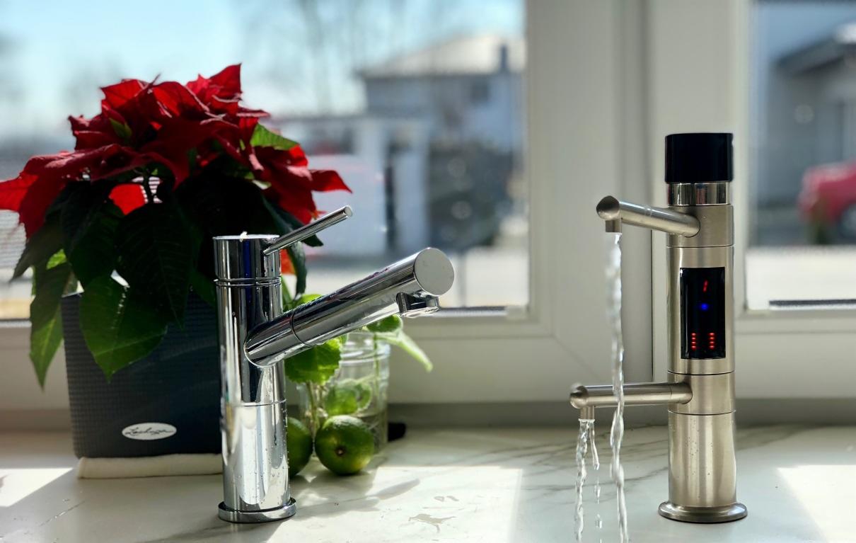 Bateria kuchenna chromowana z kranem filtrującym wodę i wyświetlaczem, obok doniczka z poinsecją i limonki w słoiku, na tle okna.