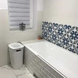 Biała wanna obudowana płytkami typu metro, nad nią dekoracyjne płytki w kwiatowy wzór. Widoczny kosz na pranie i okno z żaluzjami w łazience po remoncie w Nadarzynie.