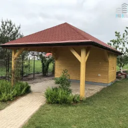 Drewniana altana z czerwoną dachówką, widoczna z zewnątrz, otoczona zielonym trawnikiem i ścieżką z kostki, z logo firmy budowlanej w prawym górnym rogu.