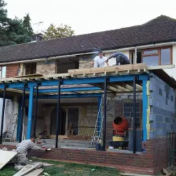 Rozbudowa domu jednorodzinnego: montaż stalowej konstrukcji nośnej, murowanie ścian z bloczków i cegły, widoczna betoniarka i narzędzia budowlane.