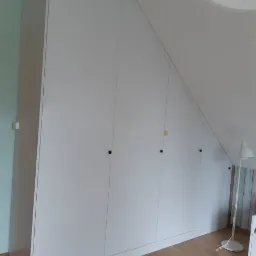 Biała szafa wnękowa z czterema drzwiami dopasowana do skosu poddasza, czarne okrągłe uchwyty, drewniana podłoga, lampa stojąca w tle.