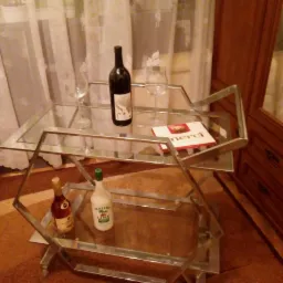 Metalowy wózek barowy z dwoma szklanymi półkami, na których stoją butelki alkoholu, kieliszki i pudełko czekoladek Merci, całość w domowym wnętrzu z drewnianą szafką w tle.