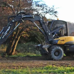 Żółto-czarna koparka kołowa Mecalac MTX w trakcie pracy na polu, z widocznym wykopem i drzewami w tle