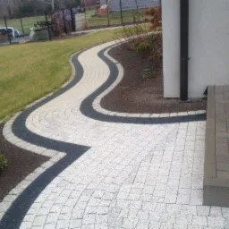 Kręta ścieżka z jasnej kostki brukowej z ciemnymi obrzeżami, prowadząca przez trawnik i obok budynku, widok z góry.