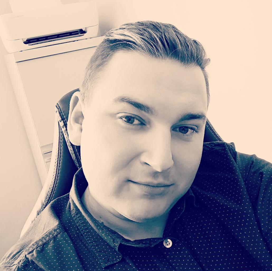 Portret mężczyzny w koszuli w kropki siedzącego na skórzanym krześle, z klimatyzatorem i szafką w tle.