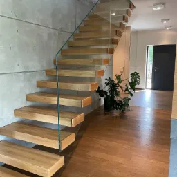 Nowoczesne schody z drewnianymi stopniami i szklaną balustradą, zamontowane przy surowej betonowej ścianie, z widokiem na korytarz z drewnianą podłogą i czarnymi drzwiami.