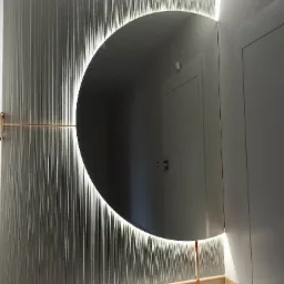 Okrągłe lustro z podświetleniem LED na ścianie z dekoracyjną tapetą w pionowe prążki, odbijające drzwi w głębi pomieszczenia.