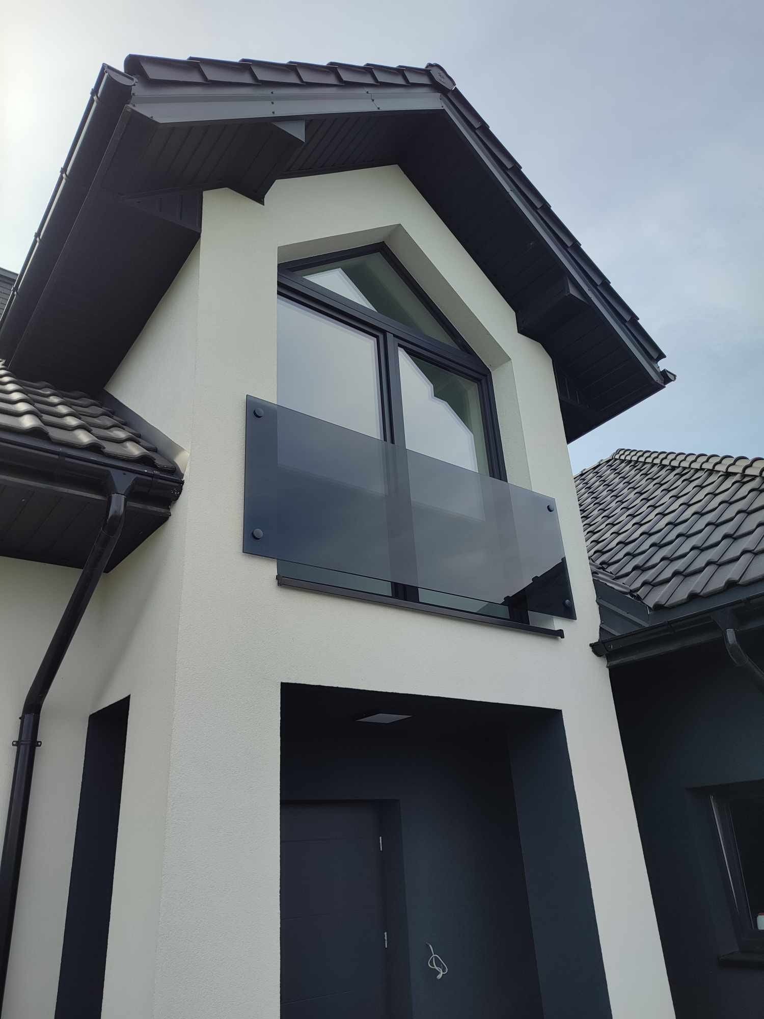 Nowoczesny dom z balkonem zabezpieczonym szklaną balustradą, widok z dołu na elewację z ciemną stolarką okienną i grafitowym dachem.