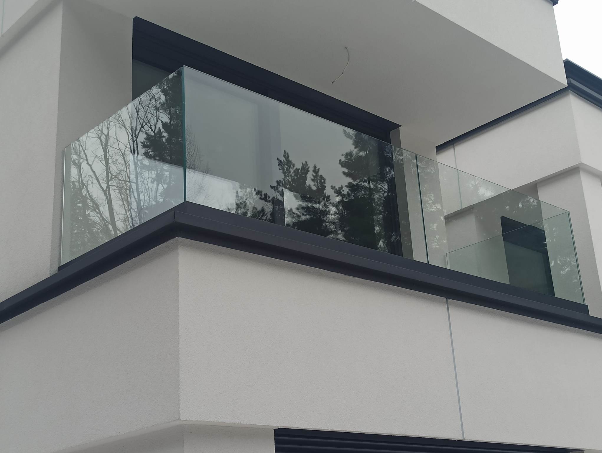 Nowoczesny balkon z przezroczystą balustradą, czarne obramowanie, widok z dołu na fragment budynku, odbicie drzew w szkle.
