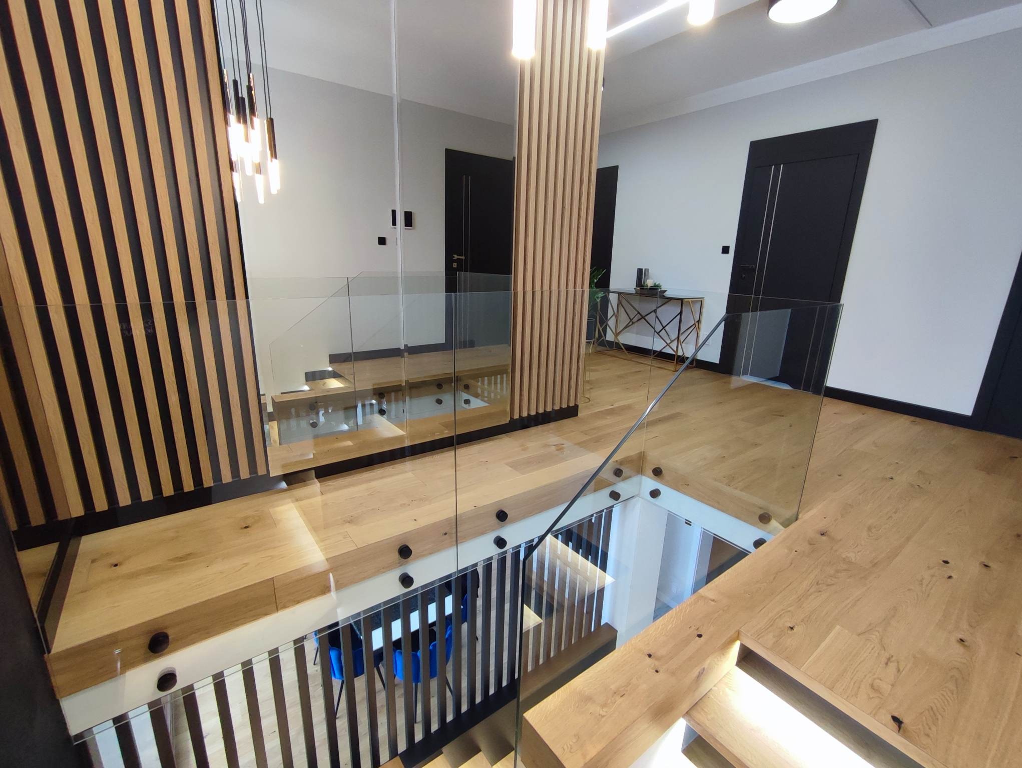 Nowoczesne wnętrze domu z drewnianymi panelami i szklaną balustradą schodową, widok z góry na schody z czarnymi poręczami.
