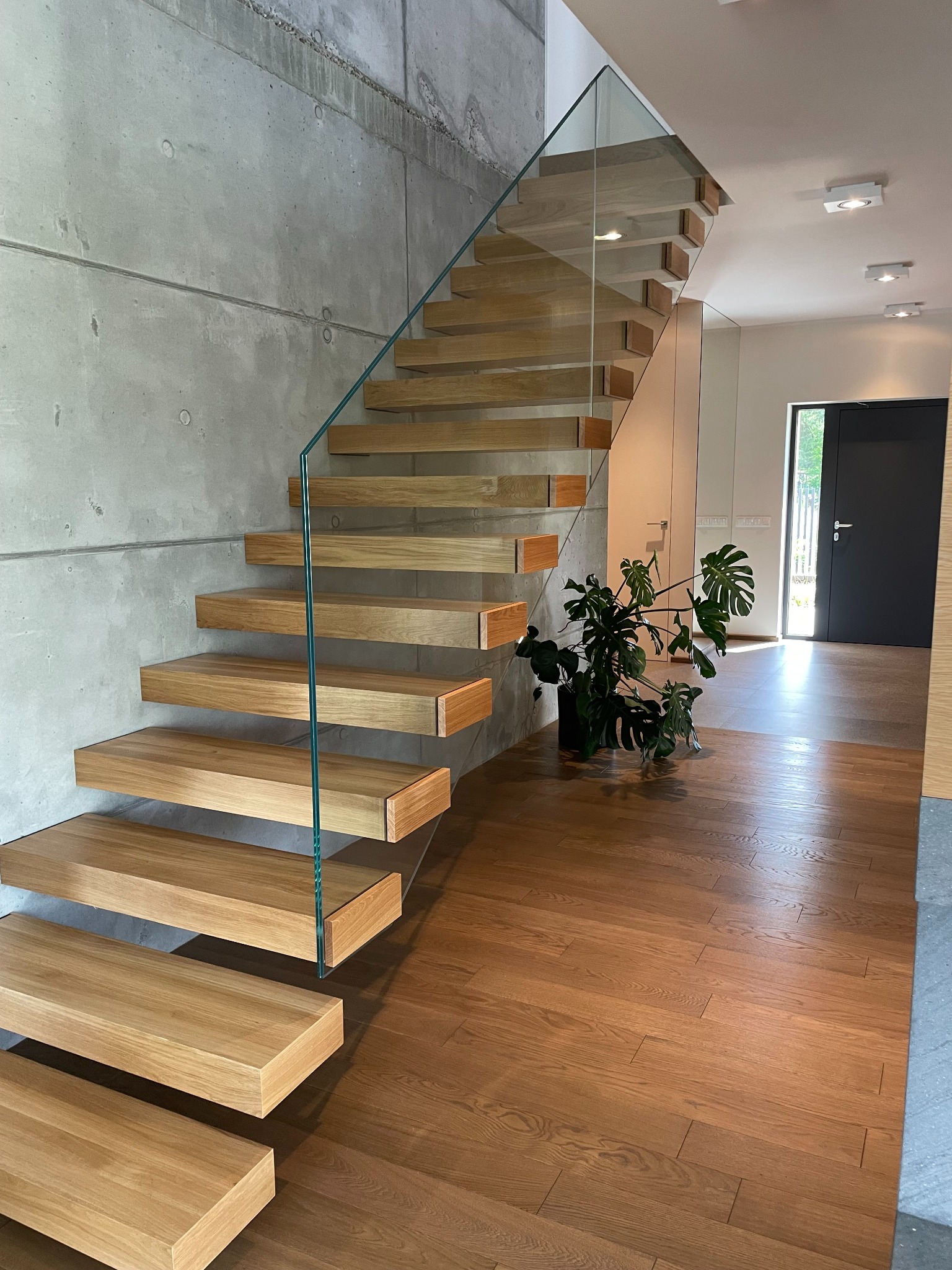 Nowoczesne schody z drewnianymi stopniami i szklaną balustradą, zamontowane przy surowej betonowej ścianie, z widokiem na korytarz z drewnianą podłogą i czarnymi drzwiami.