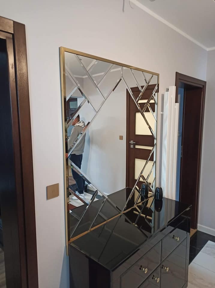 Kwadratowe lustro dekoracyjne z geometrycznym wzorem z fazowanych szklanych elementów w złotej ramie, zawieszone nad czarną komodą w przedpokoju. W lustrze odbija się osoba robiąca zdjęcie.
