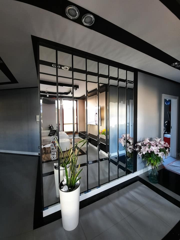 Szklana ścianka działowa z czarnymi ramami oddzielająca przestrzeń, odbijająca wnętrze salonu z białymi meblami i roślinami w donicach, widoczna z perspektywy korytarza z kwiatami w wazonie.
