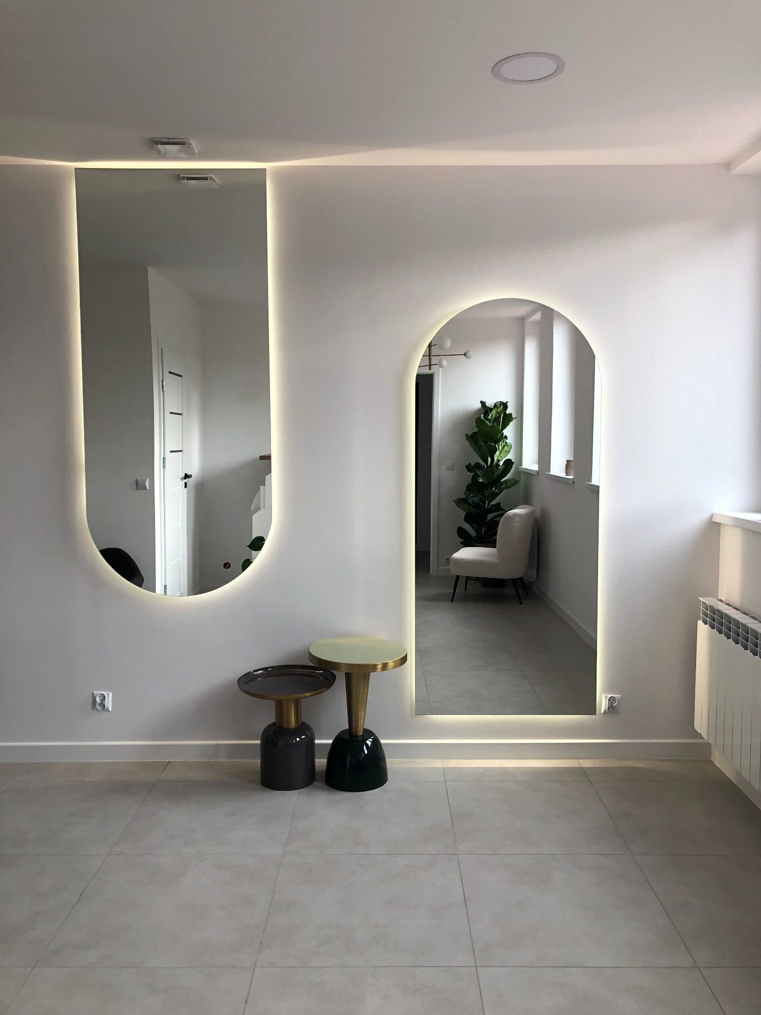 Dwa podświetlane lustra o zaokrąglonych kształtach na białej ścianie, odzwierciedlające wnętrze mieszkania z rośliną i krzesłem, uzupełnione dwoma stolikami pomocniczymi o różnych kolorach podstaw.