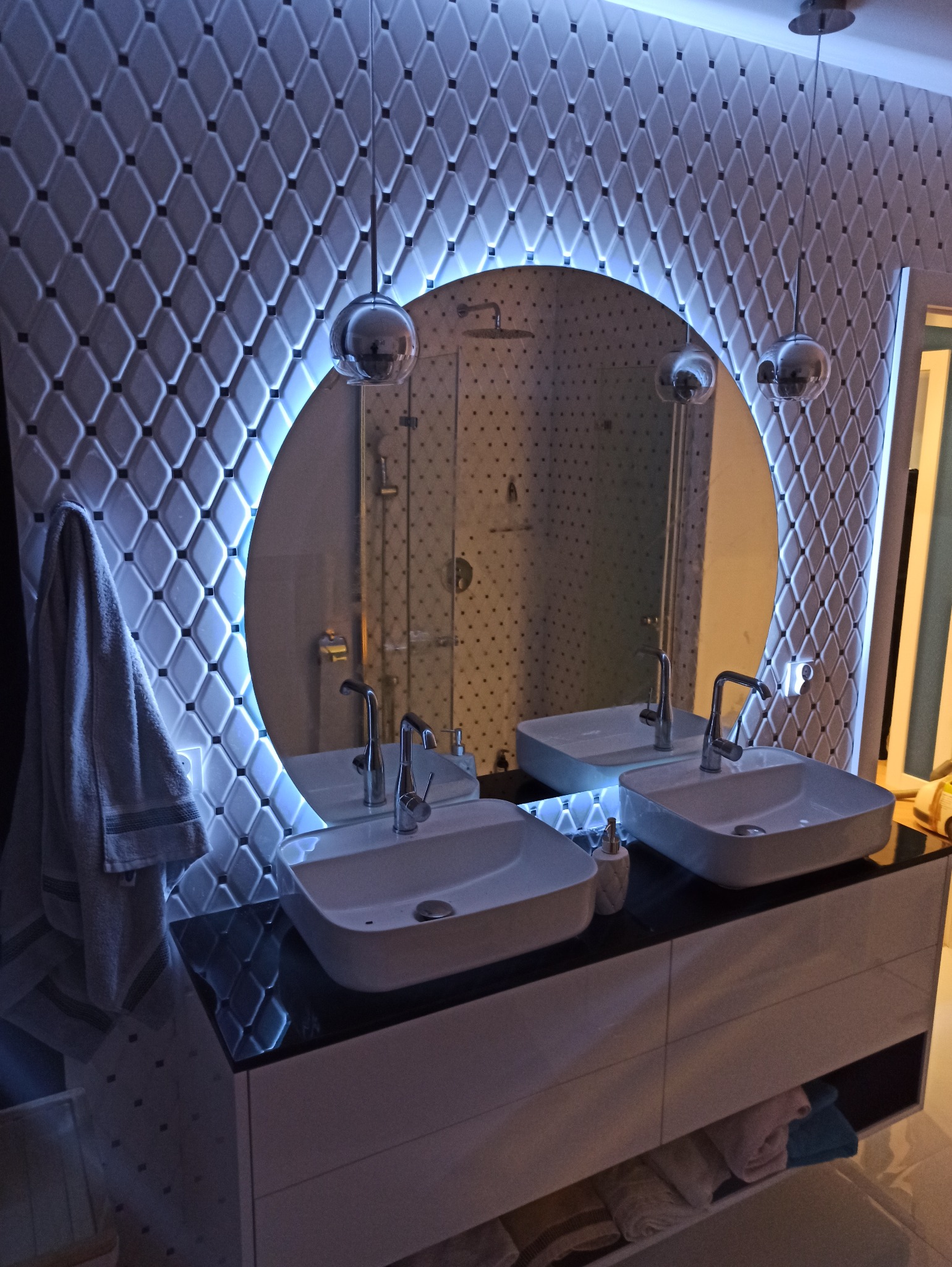 Nowoczesna łazienka z dwoma umywalkami nablatowymi, okrągłym lustrem z podświetleniem LED i dekoracyjną ścianą w romby, oświetlona wiszącymi lampami kulami.