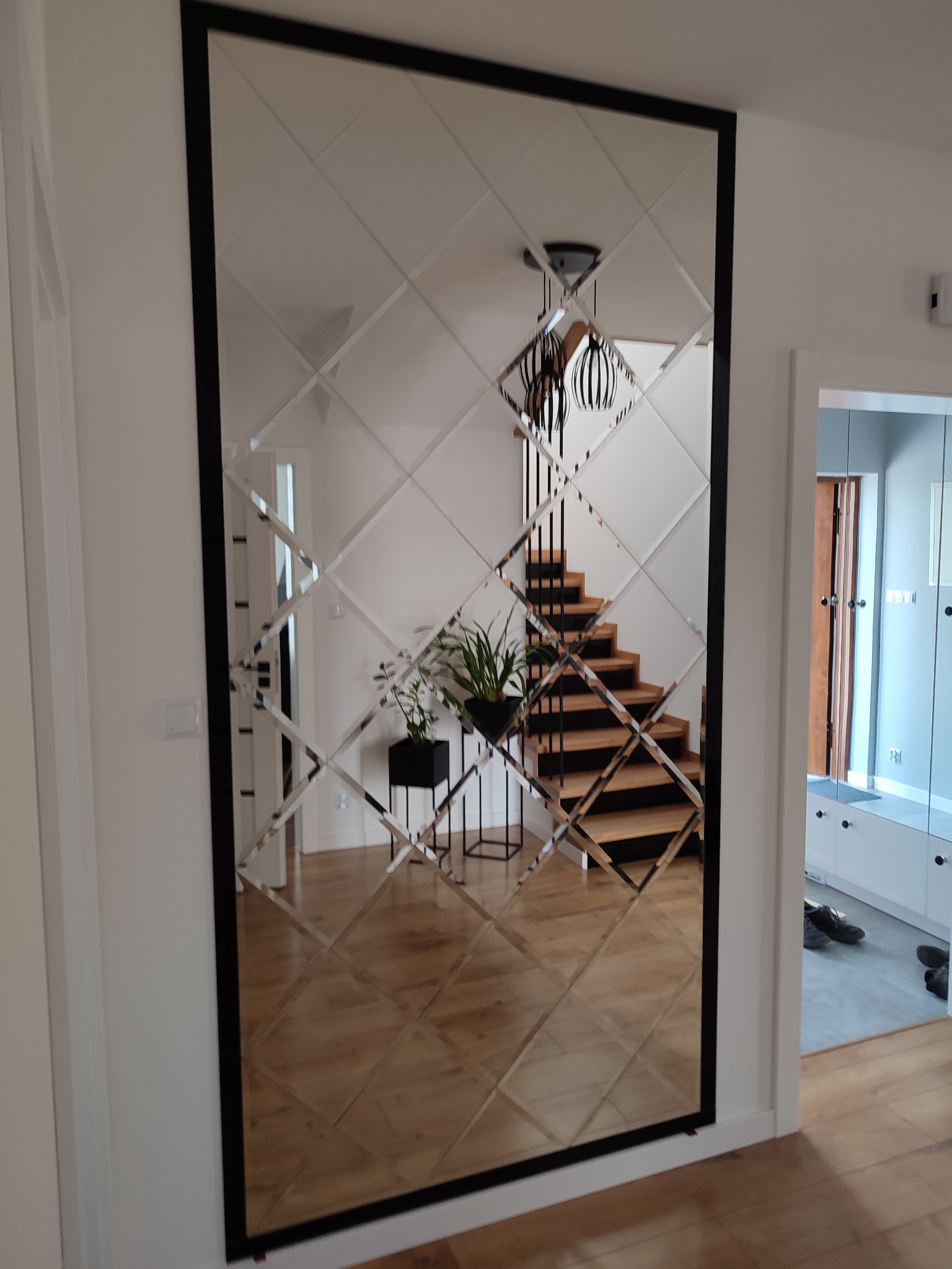 Dekoracyjne lustro ścienne z czarną ramą, składające się z romboidalnych elementów, odbijające schody z drewnianymi stopniami i nowoczesną lampę wiszącą.