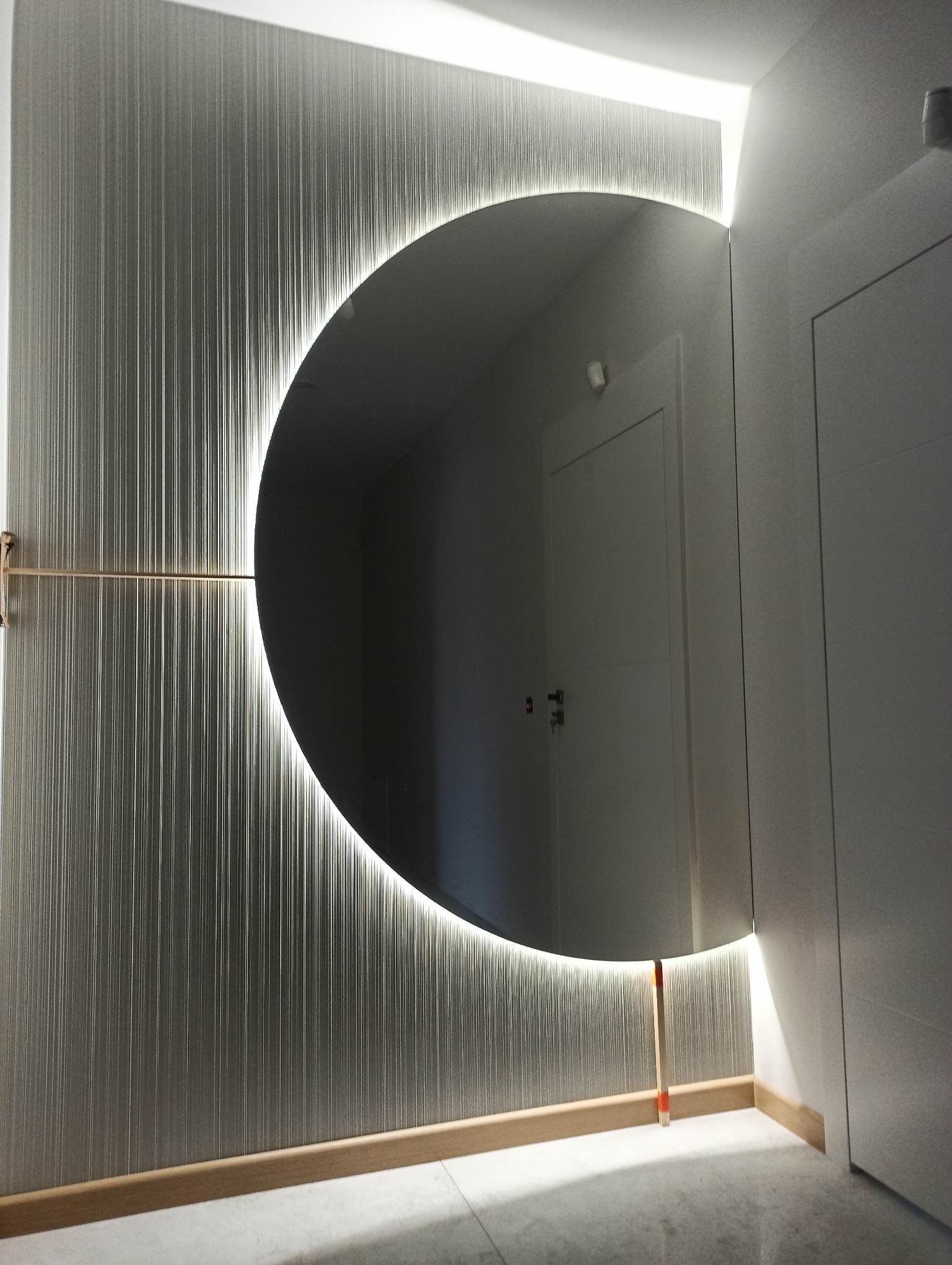 Okrągłe lustro z podświetleniem LED na ścianie z dekoracyjną tapetą w pionowe prążki, odbijające drzwi w głębi pomieszczenia.