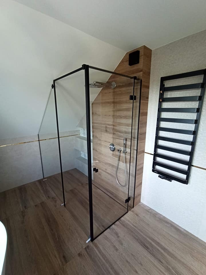 Nowoczesna kabina prysznicowa ze szklanymi ściankami i czarnymi profilami, dopasowana do skośnego sufitu w łazience z drewnopodobnymi płytkami.