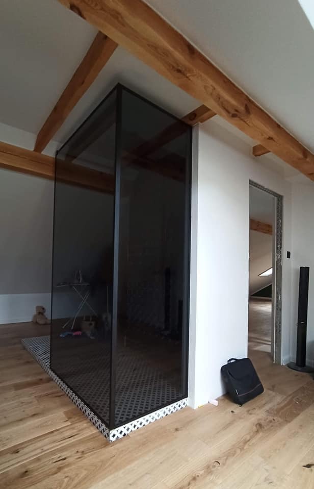 Szklana konstrukcja narożna z czarną ramą, oddzielająca przestrzeń na poddaszu z drewnianymi belkami stropowymi i podłogą z jasnego drewna.