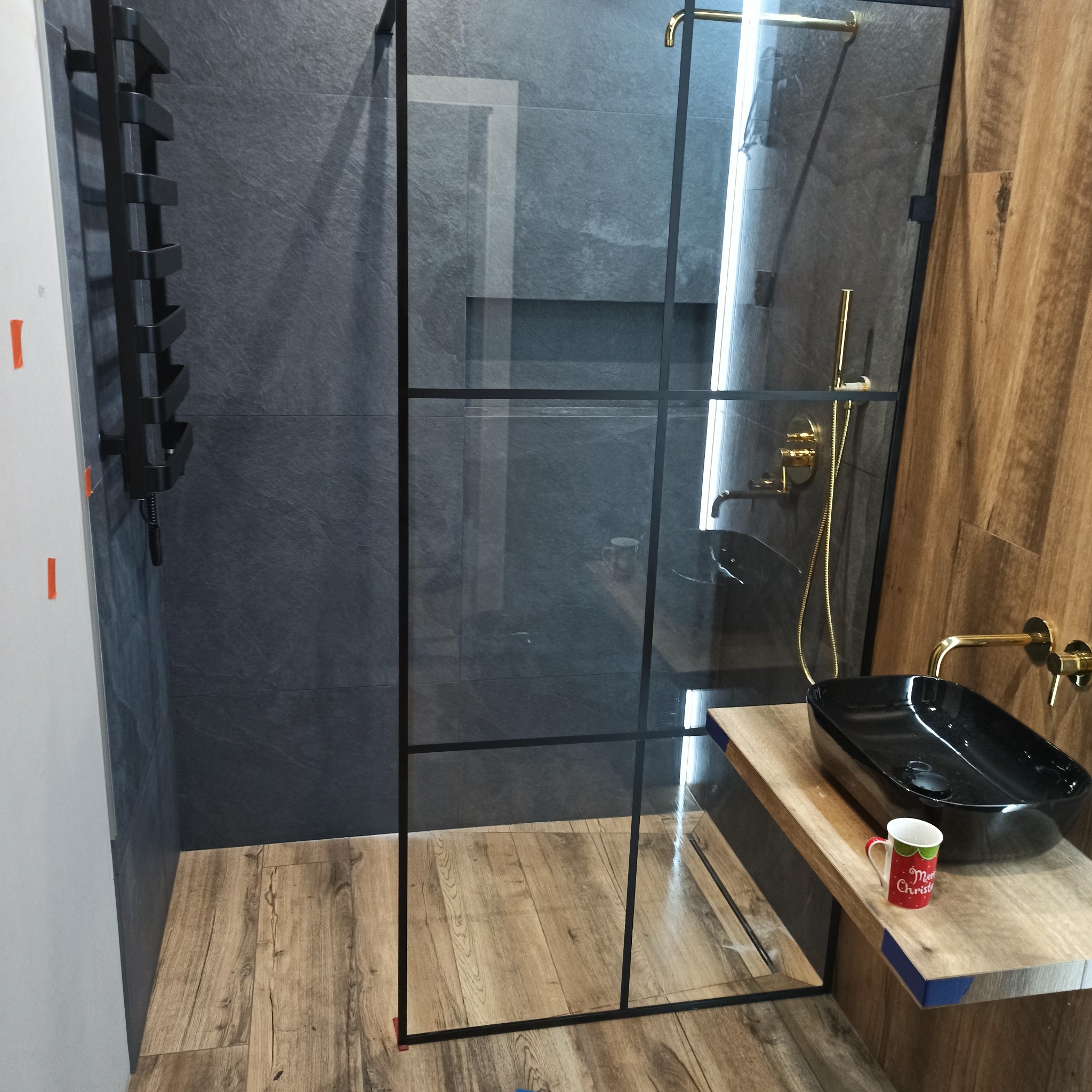 Nowoczesna łazienka z kabiną prysznicową typu walk-in z czarnymi profilami, złotą armaturą i czarną umywalką na drewnianym blacie, uzupełniona grzejnikiem drabinkowym i świątecznym kubkiem.