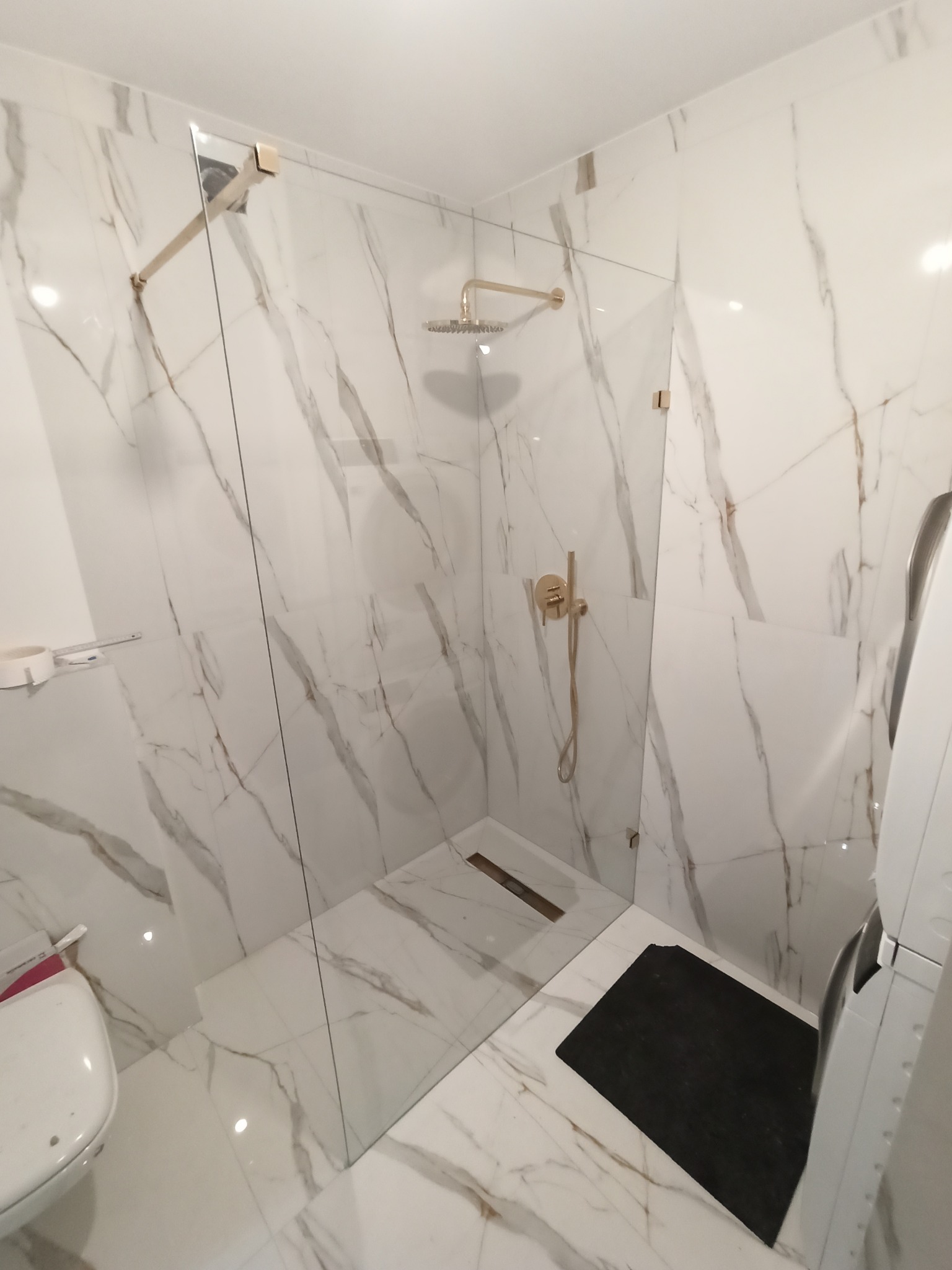 Nowoczesna kabina prysznicowa ze szklaną ścianą i złotą armaturą, marmurowe płytki na ścianach i podłodze, czarny dywanik łazienkowy, widoczna toaleta i pralka.