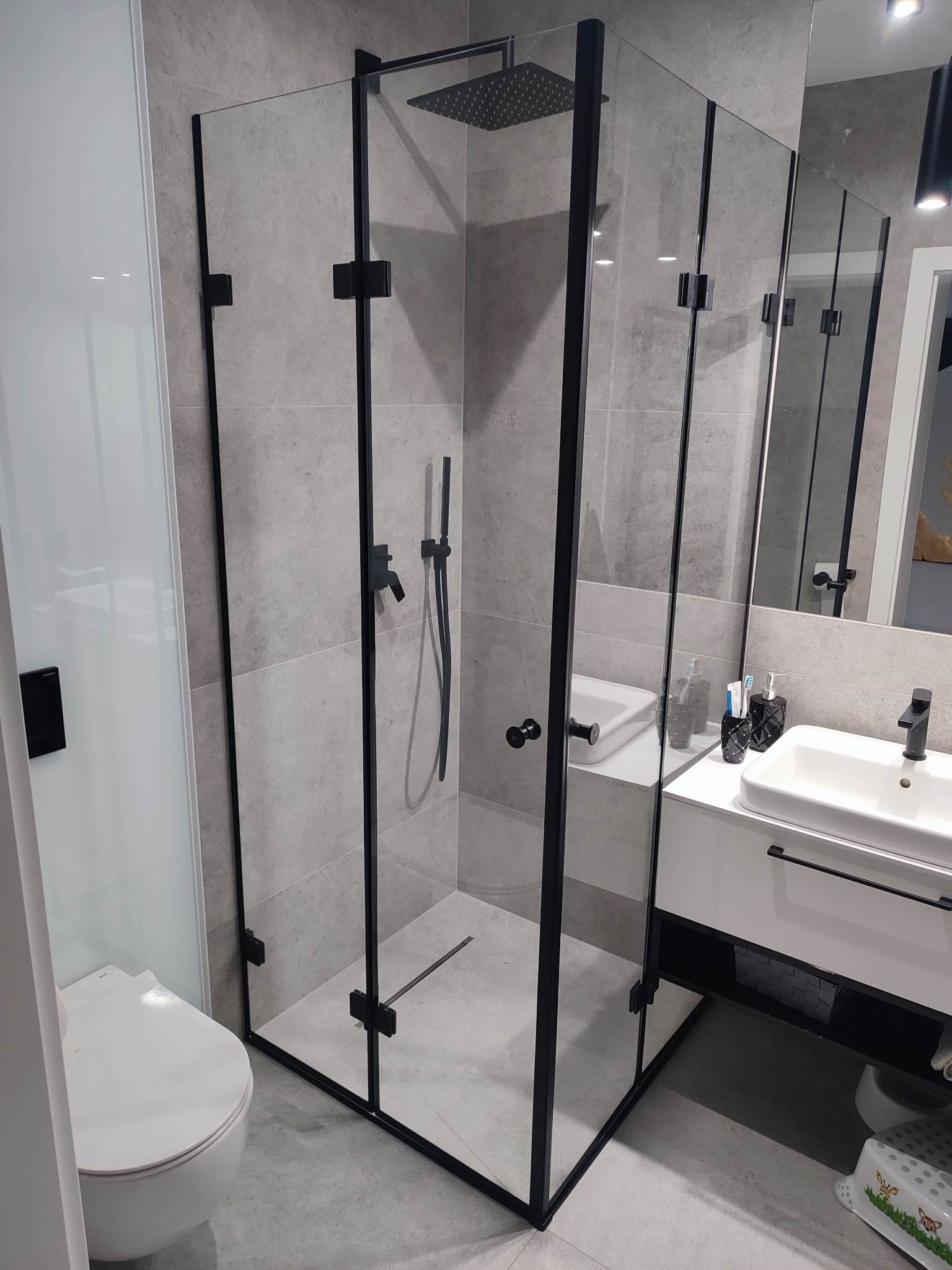 Nowoczesna kabina prysznicowa z czarnymi profilami i deszczownicą w łazience wykończonej szarymi płytkami, widoczna umywalka z czarną armaturą i toaleta.