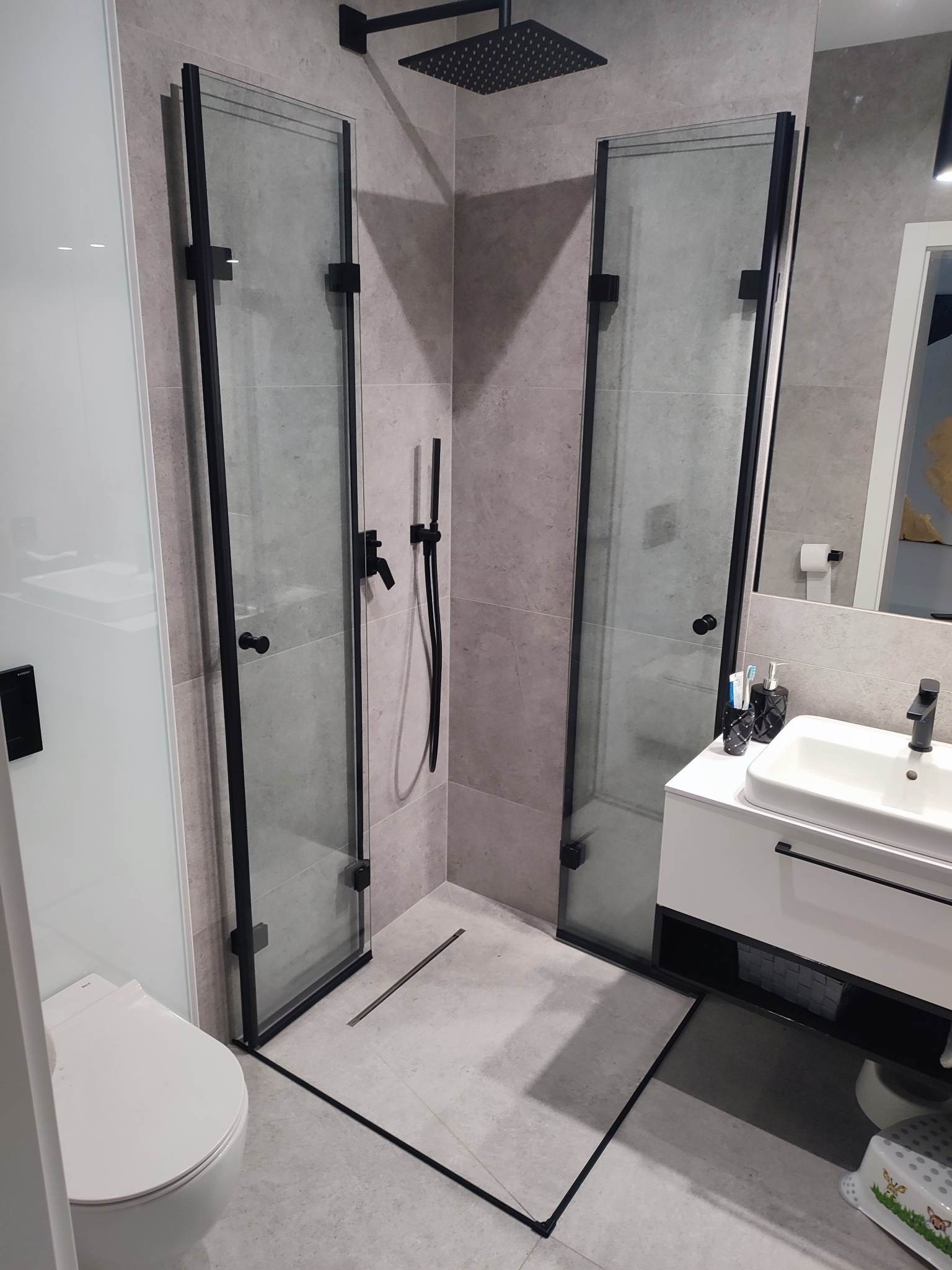 Nowoczesna łazienka z kabiną prysznicową typu walk-in, wykończoną czarnymi profilami i armaturą, z odpływem liniowym w podłodze i umywalką nablatową.