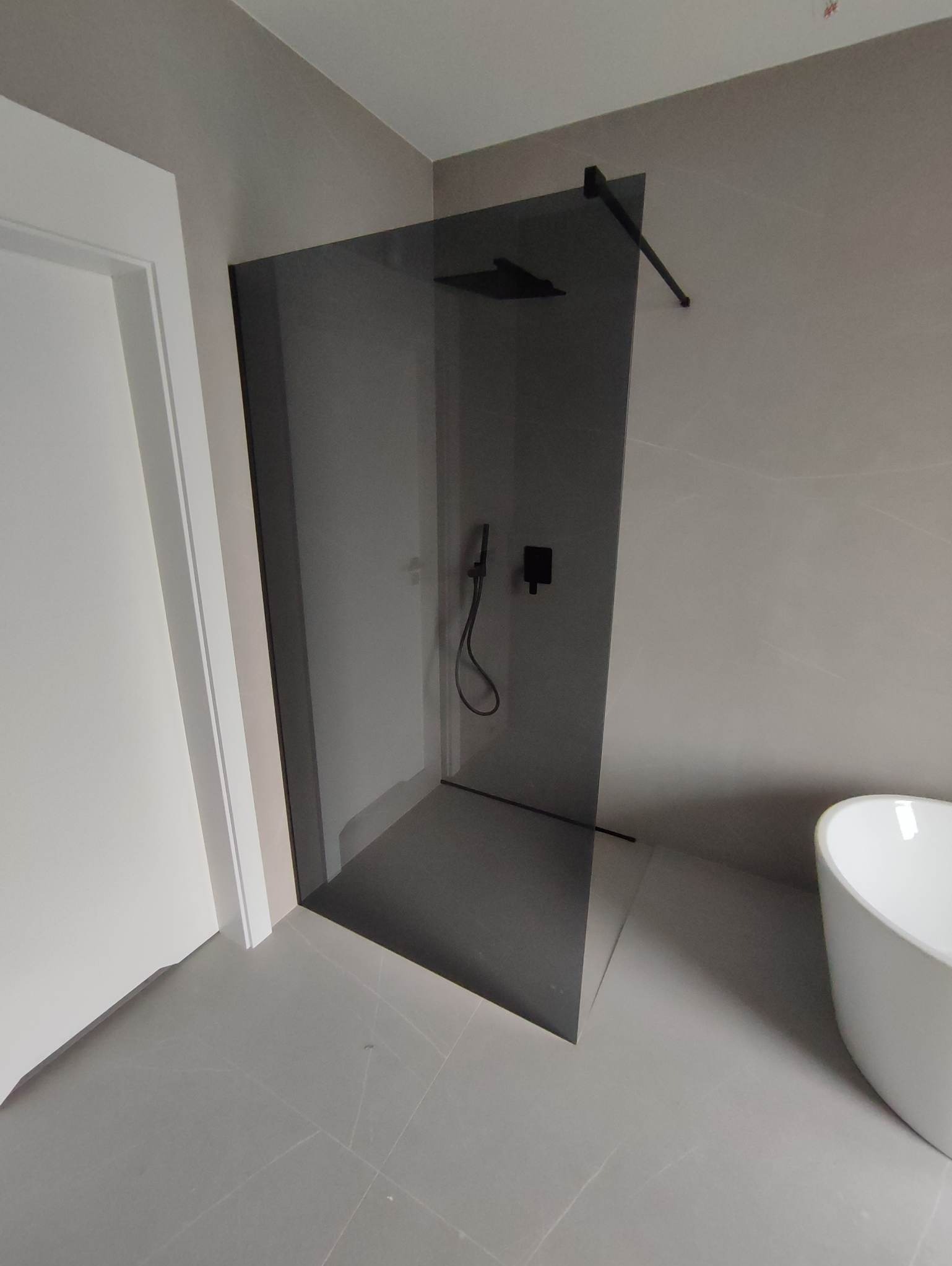 Nowoczesna kabina prysznicowa z przyciemnianego szkła, wyposażona w czarną armaturę, w minimalistycznej łazience z szarą glazurą i białą wanną wolnostojącą.