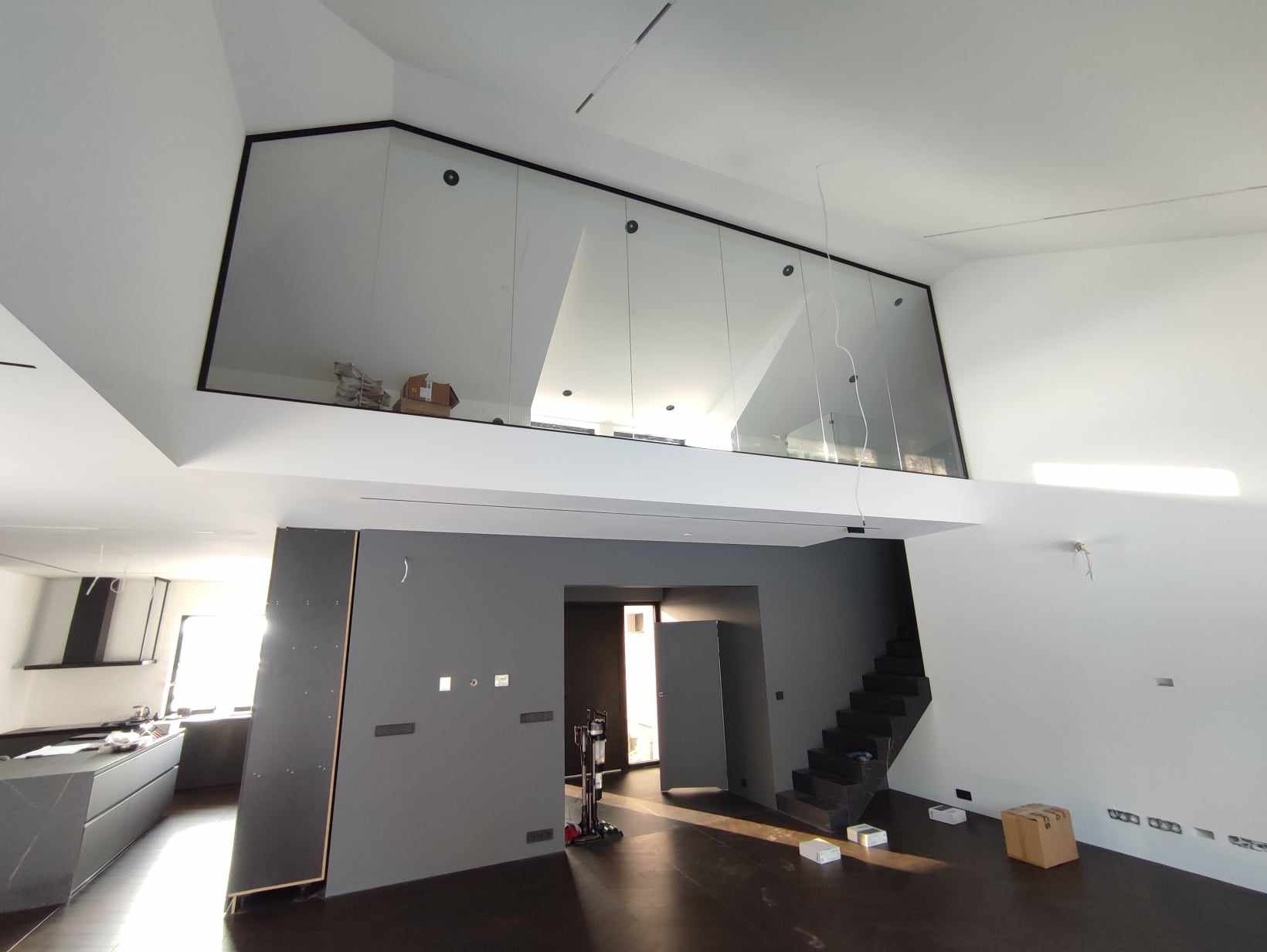 Nowoczesne wnętrze z antresolą zabezpieczoną szklaną balustradą w czarnej ramie, widok na otwartą kuchnię i schody.