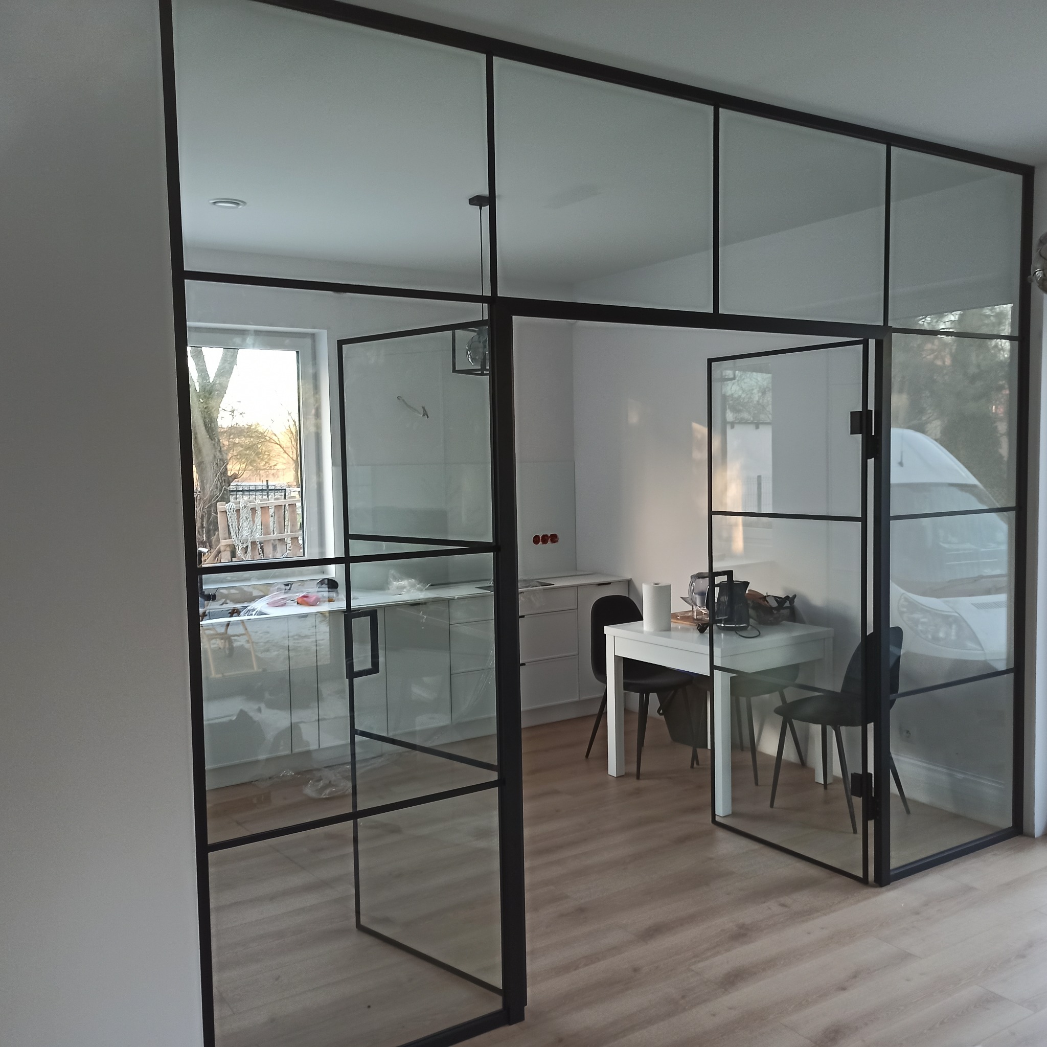 Szklana ścianka działowa z czarnymi profilami oddzielająca kuchnię od reszty mieszkania, widoczny stół z krzesłami i okno z widokiem na zewnątrz.