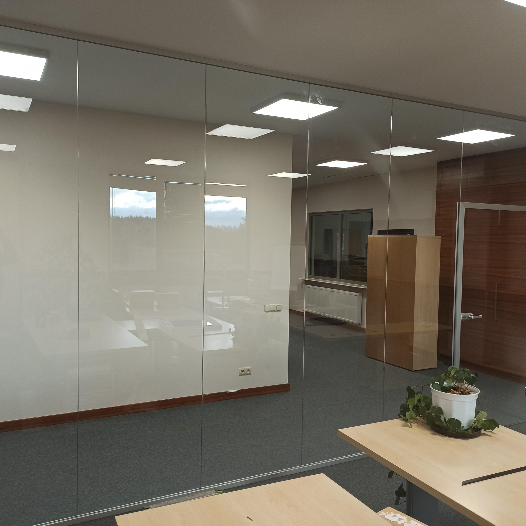 Szklane ścianki działowe w biurze, widoczne biurka, krzesła i rośliny doniczkowe, światło odbija się od powierzchni szkła.