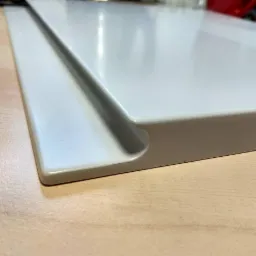 Fragment białego, lakierowanego frontu meblowego z frezowanym uchwytem krawędziowym, leżącego na blacie biurka.