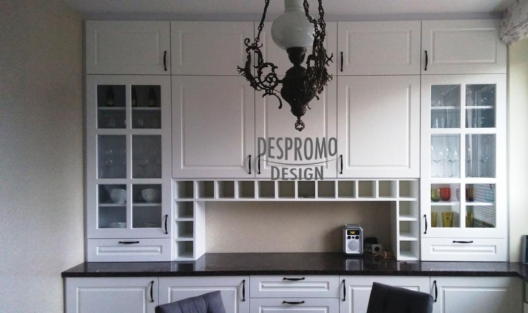 Biała zabudowa kuchenna z górnymi szafkami z przeszklonymi frontami, wbudowanymi półkami na wino i czarnym blatem, oświetlona ozdobnym, wiszącym żyrandolem z kutego żelaza.