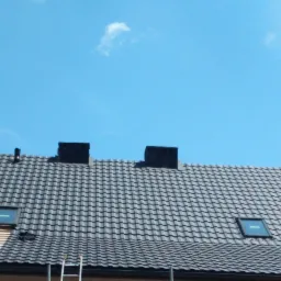 Szary dach z dachówki ceramicznej z dwoma kominami i oknami dachowymi, fragment rusztowania i drabina oparta o budynek.