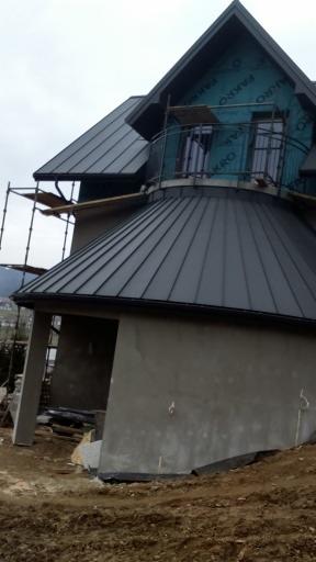 Dom w trakcie budowy z ciemnym, metalowym dachem, widoczne rusztowania i folia ochronna na poddaszu, teren wokół budynku częściowo rozkopany.