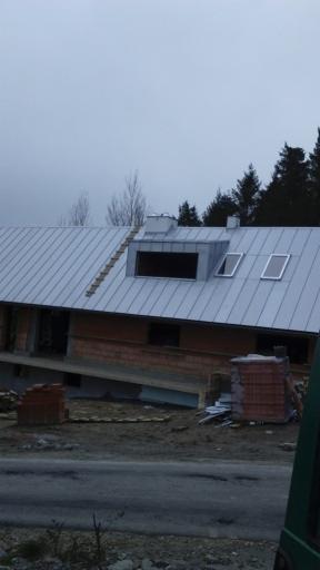 Dom w trakcie budowy z metalowym dachem, widoczne okna dachowe i komin, sterty cegieł na zewnątrz, pochmurne niebo w tle.